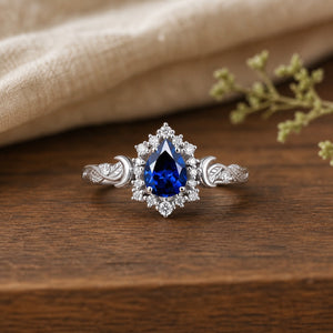 Pear-cut royal-blue sapphire leaf-and-moon halo engagement ring Luna on a warm walnut tabletop