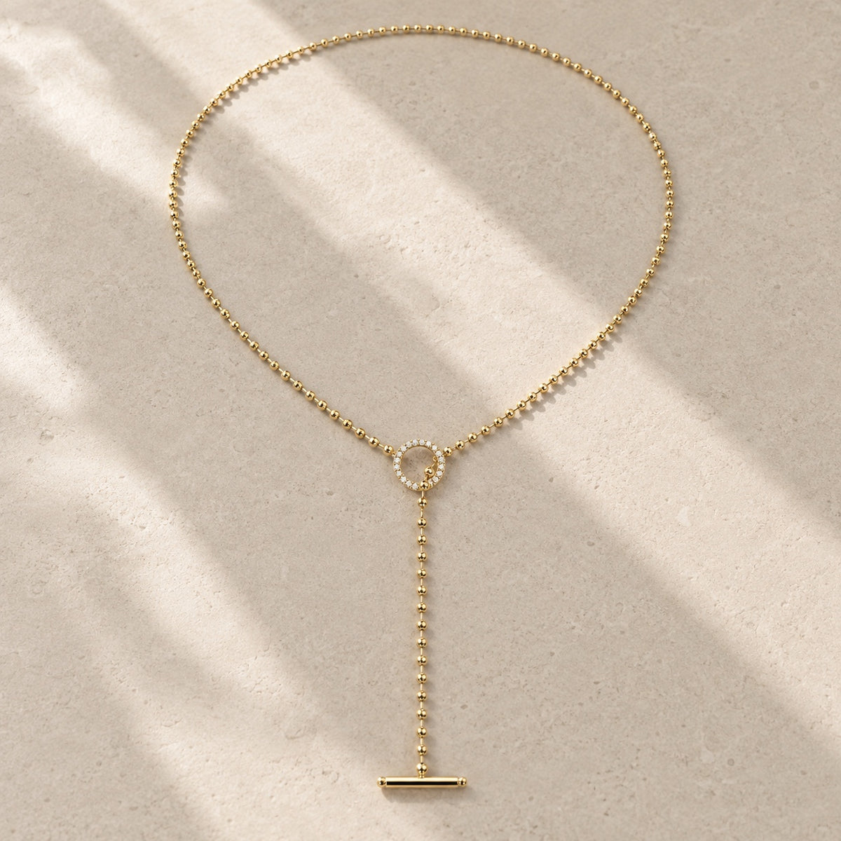 Gold beaded lariat toggle necklace with moissanite ring and bar pendant on pale stone