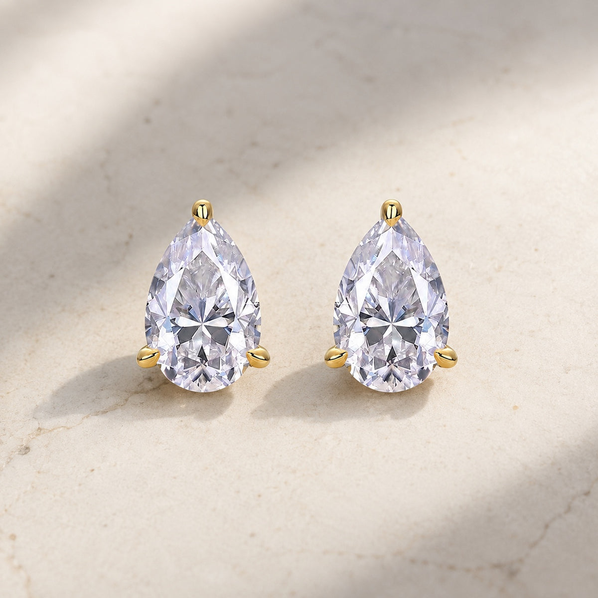 Pair of pear-shaped clear moissanite stud earrings with gold prongs on pale stone